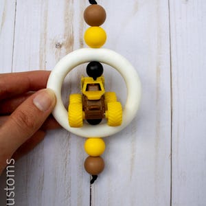 K&ouml;nnte beinhalten: Ein Baby-Bei&szlig;spielzeug mit einem gelb-braunen Spielzeug-LKW in einem wei&szlig;en Ring. Das Bei&szlig;spielzeug hat gelbe, braune und schwarze Perlen und eine schwarze Schnur. Der Text "907 Customs" ist auf der linken Seite sichtbar.