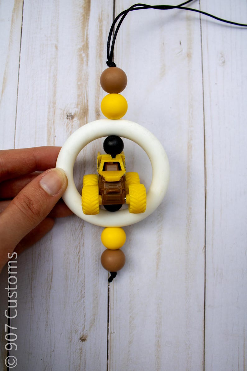 K&ouml;nnte beinhalten: Ein Baby-Bei&szlig;spielzeug mit einem gelb-braunen Spielzeug-LKW in einem wei&szlig;en Ring. Das Bei&szlig;spielzeug hat gelbe, braune und schwarze Perlen und eine schwarze Schnur. Der Text "907 Customs" ist auf der linken Seite sichtbar.