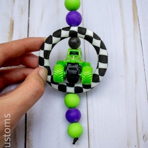 May include: A green and black monster truck toy within a black and white chequered ring, suspended by a black cord with purple and green beads. The image includes the text "907 Customs".
