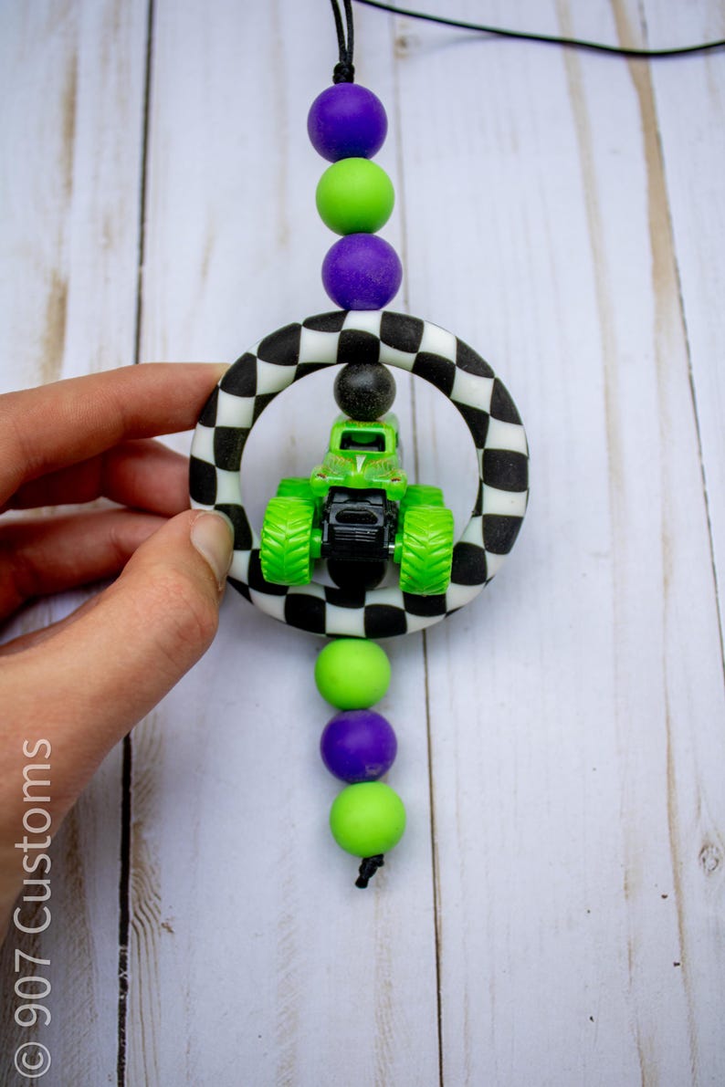 May include: A green and black monster truck toy within a black and white chequered ring, suspended by a black cord with purple and green beads. The image includes the text "907 Customs".