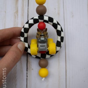 May include: A car-themed baby toy. The toy features a small race car inside a black and white chequered ring, with yellow, brown, and red accents. The toy is suspended by a white string.