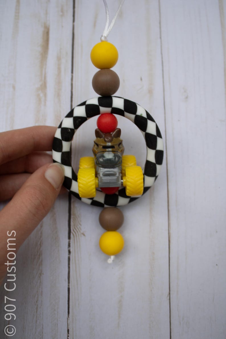 May include: A car-themed baby toy. The toy features a small race car inside a black and white chequered ring, with yellow, brown, and red accents. The toy is suspended by a white string.