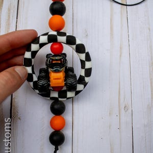 K&ouml;nnte beinhalten: Ein kleiner Monster-Truck in Orange und Schwarz wird von einem schwarz-wei&szlig; karierten Ring umrahmt. Das Spielzeug wird von einer schwarzen Schnur mit orangefarbenen und schwarzen Perlen gehalten. Der Text "907 Customs" ist sichtbar.
