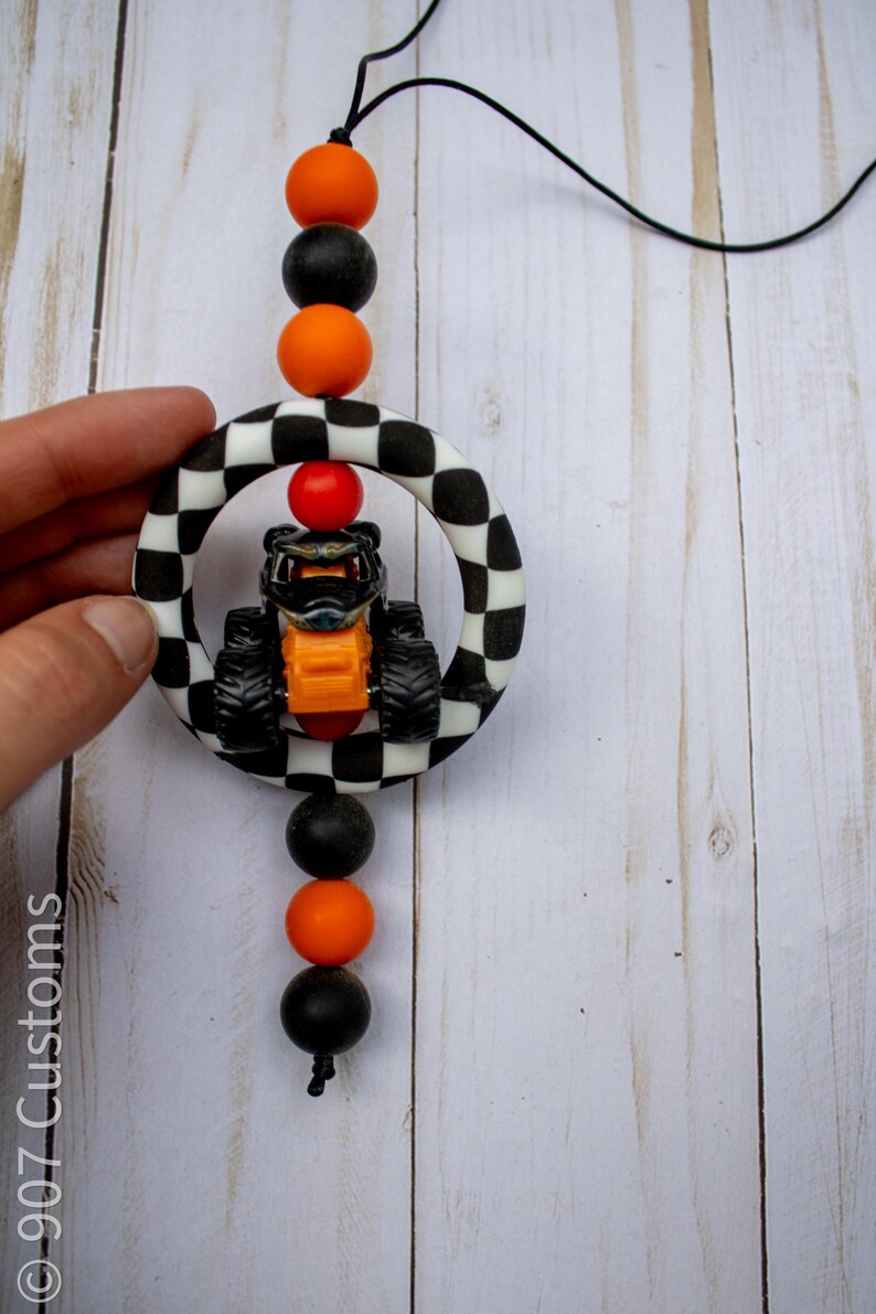 K&ouml;nnte beinhalten: Ein kleiner Monster-Truck in Orange und Schwarz wird von einem schwarz-wei&szlig; karierten Ring umrahmt. Das Spielzeug wird von einer schwarzen Schnur mit orangefarbenen und schwarzen Perlen gehalten. Der Text "907 Customs" ist sichtbar.