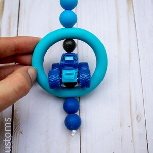 May include: A blue and turquoise teething toy featuring a small blue tractor inside a turquoise ring. The toy is strung with blue beads and a white cord. The toy is held by a hand.