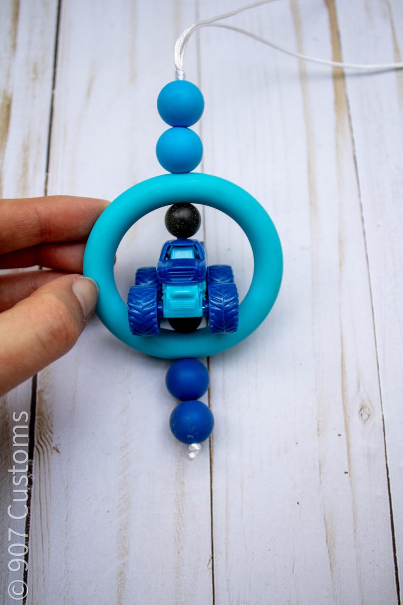 May include: A blue and turquoise teething toy featuring a small blue tractor inside a turquoise ring. The toy is strung with blue beads and a white cord. The toy is held by a hand.