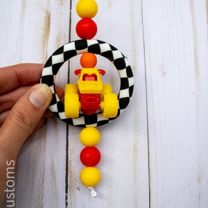 K&ouml;nnte beinhalten: Ein farbenfrohes Babyspielzeug mit einem gelb-roten Rennwagen in einem schwarz-wei&szlig; karierten Ring. Das Spielzeug ist mit roten und gelben Perlen verziert, die auf einer wei&szlig;en Schnur aufgef&auml;delt sind. Das Design ist verspielt und lebendig.