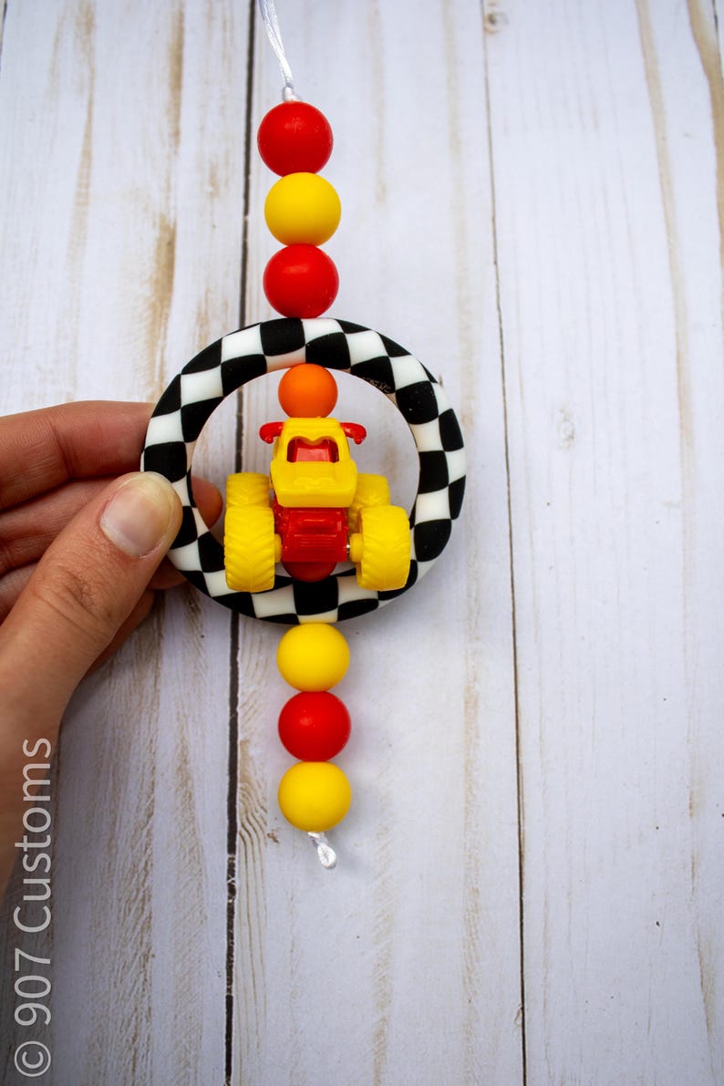 K&ouml;nnte beinhalten: Ein farbenfrohes Babyspielzeug mit einem gelb-roten Rennwagen in einem schwarz-wei&szlig; karierten Ring. Das Spielzeug ist mit roten und gelben Perlen verziert, die auf einer wei&szlig;en Schnur aufgef&auml;delt sind. Das Design ist verspielt und lebendig.