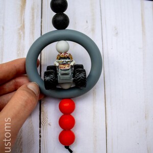 May include: A grey teething ring with a small monster truck toy inside, connected to a string with black and red beads. The text "907 Customs" is visible on the left side.