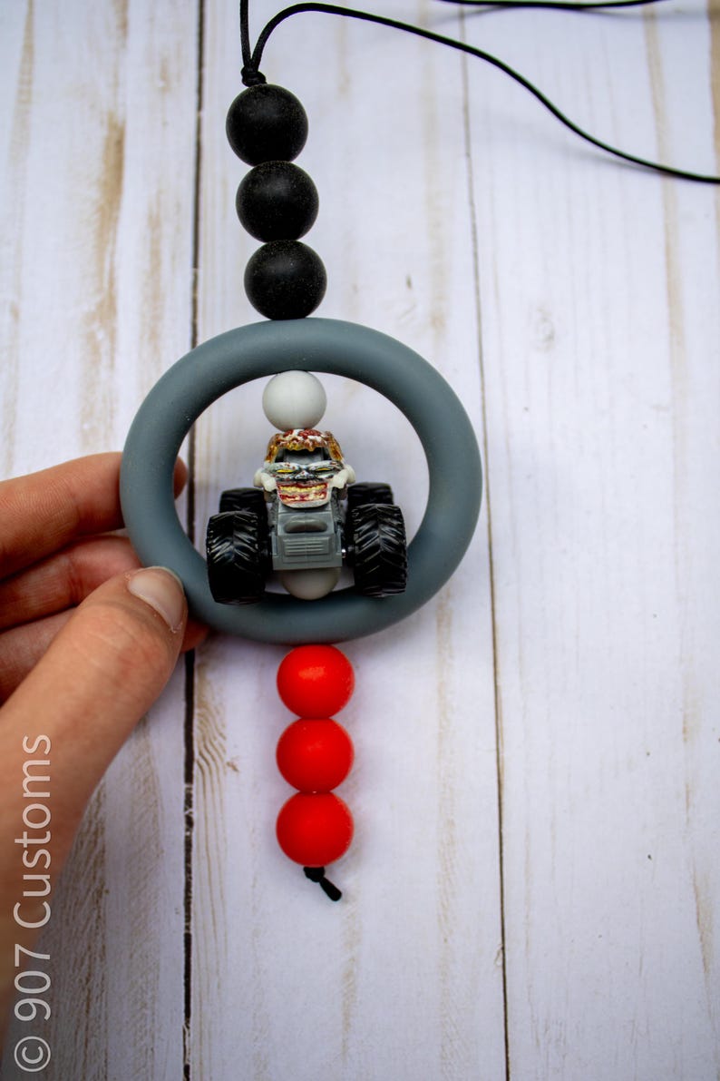 May include: A grey teething ring with a small monster truck toy inside, connected to a string with black and red beads. The text "907 Customs" is visible on the left side.