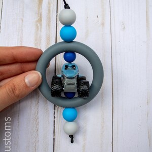 K&ouml;nnte beinhalten: Ein blaues Monstertruck-Spielzeug befindet sich in einem grauen Silikonring, der an einer schwarzen Schnur h&auml;ngt. Die Schnur ist mit grauen und blauen Perlen aufgef&auml;delt. Der Hintergrund ist ein helles Holzmaserungsmuster.