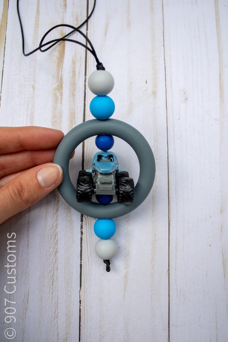 K&ouml;nnte beinhalten: Ein blaues Monstertruck-Spielzeug befindet sich in einem grauen Silikonring, der an einer schwarzen Schnur h&auml;ngt. Die Schnur ist mit grauen und blauen Perlen aufgef&auml;delt. Der Hintergrund ist ein helles Holzmaserungsmuster.