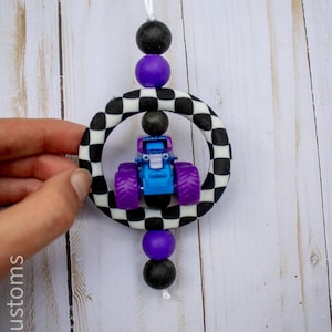 May include: A blue and purple monster truck toy is suspended within a black and white chequered ring, accented by black and purple beads. The toy is attached to a white string, creating a playful, decorative item.