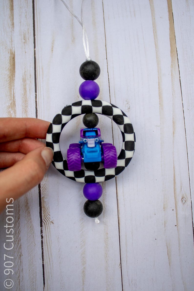 May include: A blue and purple monster truck toy is suspended within a black and white chequered ring, accented by black and purple beads. The toy is attached to a white string, creating a playful, decorative item.