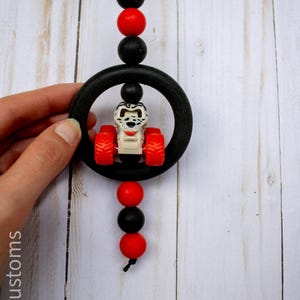 K&ouml;nnte beinhalten: Ein schwarz-roter Perlen-Autoanh&auml;nger mit einem kleinen Spielzeugtraktor in der Mitte. Der Anh&auml;nger hat einen schwarzen Ring und ist an einer schwarzen Schnur aufgeh&auml;ngt. Der Traktor hat rote R&auml;der und einen wei&szlig;en K&ouml;rper.