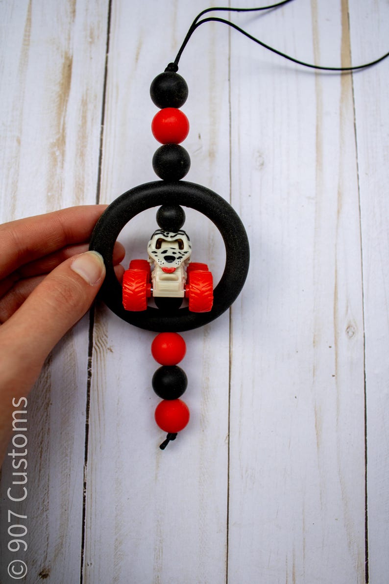 K&ouml;nnte beinhalten: Ein schwarz-roter Perlen-Autoanh&auml;nger mit einem kleinen Spielzeugtraktor in der Mitte. Der Anh&auml;nger hat einen schwarzen Ring und ist an einer schwarzen Schnur aufgeh&auml;ngt. Der Traktor hat rote R&auml;der und einen wei&szlig;en K&ouml;rper.