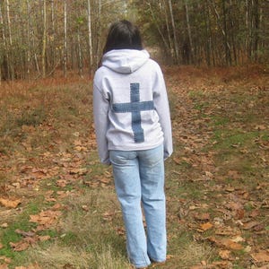 May include: A light gray hoodie with a large denim cross stitched on the back. The person is wearing light blue jeans and brown shoes. The background is a path through a forest with fallen leaves.