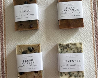 May include: Four rectangular goat milk soaps with white labels. The soaps are labeled "Cacao", "Warm Cinnamon", "Fresh Coffee", and "Lavender". The soaps are various shades of brown, beige, and green, with visible inclusions.