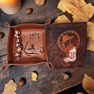 May include: A square brown leather dice tray with a fox illustration and a matching dark brown fabric dice bag featuring an embroidered fox design. A column of seven polyhedral dice with orange and white swirling patterns sits inside the tray.