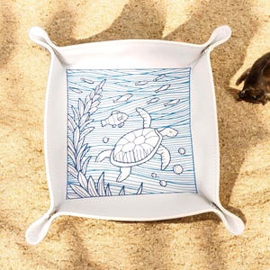 May include: White leather tray with a blue and white design of a sea turtle swimming in the ocean with fish and seaweed. The tray has a square shape with rounded corners and four handles.