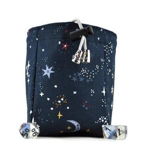 May include: A dark blue dice bag with a white and blue star pattern. The bag has a drawstring closure and is decorated with a crescent moon and stars. Two dice are visible, one with the number 6 and the other with the number 1.