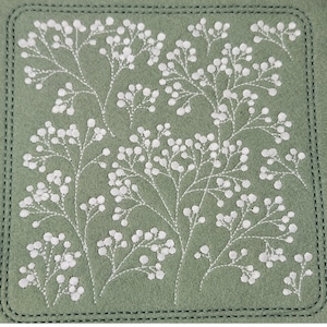 May include: A square, sage green coaster with white embroidered floral sprigs. The design features delicate white thread creating a pattern of small, round flower clusters on thin stems. The coaster has a stitched border.