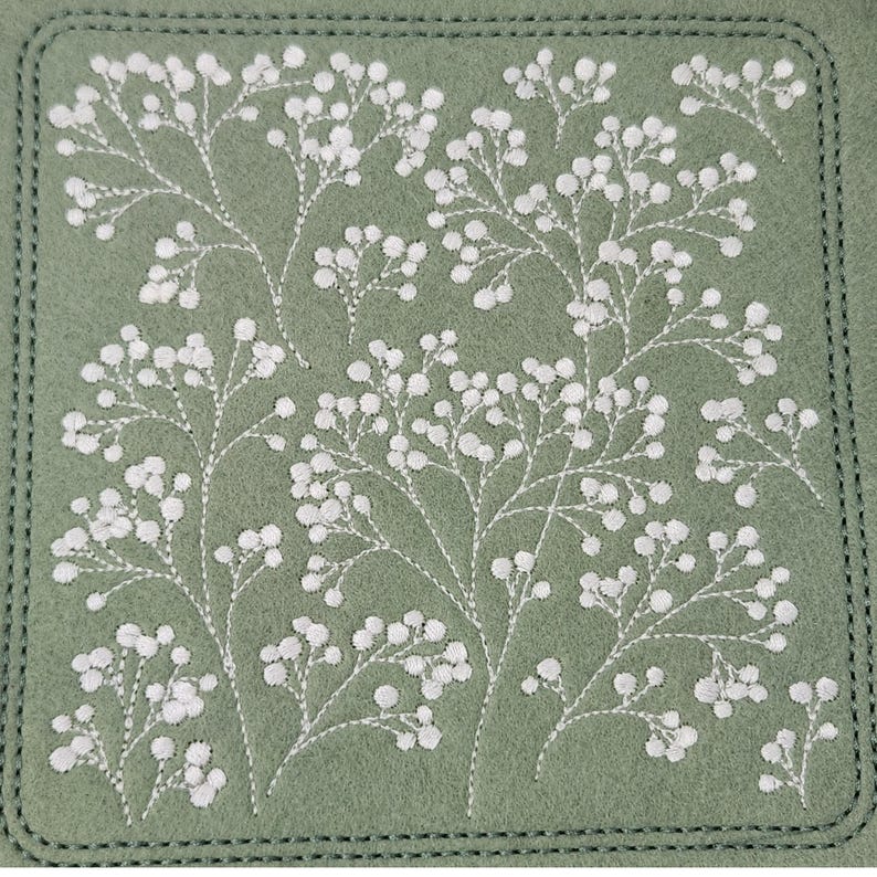 May include: A square, sage green coaster with white embroidered floral sprigs. The design features delicate white thread creating a pattern of small, round flower clusters on thin stems. The coaster has a stitched border.