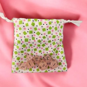 May include: A white drawstring dice bag with a pattern of green squares, pink squares, and black dots. The bag contains several light pink dice with black numbers. The background is a pink fabric.