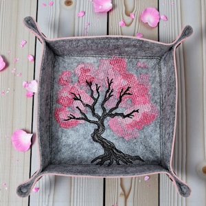 May include: A square, grey felt tray with a pink trim and handles. The tray features an embroidered cherry blossom tree with pink flowers and a black trunk. Pink flower petals are scattered around the tray.