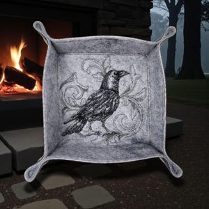 Pode incluir: Uma bandeja de feltro cinza com um desenho de corvo bordado em preto e enfeites decorativos. A bandeja tem laterais curvas e quatro alças de laço. O fundo mostra uma lareira com fogo queimando.