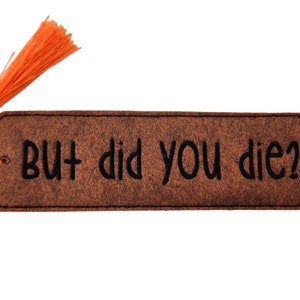 Könnte beinhalten: Braunes Filz Lesezeichen mit einem orangefarbenen Quasten und dem Text "BUT did you die?" in schwarzer Stickerei.