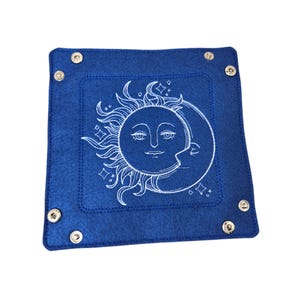 May include: A square, deep blue felt tray with a white embroidered sun and moon design. The celestial graphic features detailed faces and star accents. Silver snaps secure the corners of the tray, which is suitable for jewelry or small items.