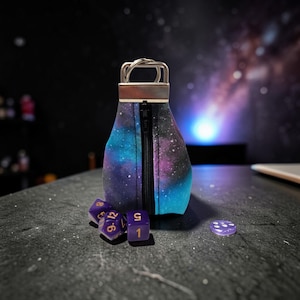 Op de afbeelding: Een kleine, cilindrische dobbelsteenzak met ritssluiting, met een levendige galaxy-print in blauw, paars en zwart. De tas heeft een zilverkleurige metalen handgreep. Verschillende paarse dobbelstenen met gouden cijfers liggen naast de tas.