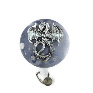 Op de afbeelding: Een ronde, blauwe badgehaspel met een zilveren drakenembleem. De badgehaspel heeft een stoffen achtergrond met zilveren stippen en een intrekbaar koord. De draak heeft gedetailleerde vleugels en een gekrulde staart.