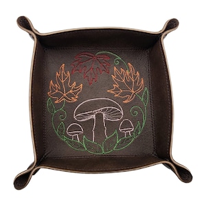 May include: A brown leather tray with a stitched design of mushrooms and fall leaves in orange, red, and green. The tray has a square shape with curved edges and folded corners.