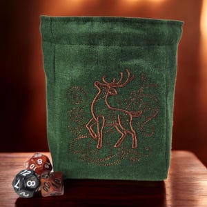 Puede incluir: Bolsa de dados de tela verde con un diseño de ciervo bordado en marrón. La bolsa está sobre una superficie de madera con tres dados a su lado.