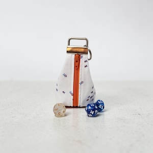Può includere: Un sacchetto per dadi in tessuto bianco con un motivo a pesci blu e arancioni, una cerniera arancione e un portachiavi in metallo. Due dadi blu e un dado trasparente sono accanto al sacchetto. Il sacchetto è progettato per contenere dadi per giochi da tavolo.