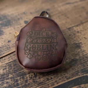 May include: A dark brown, rounded, zippered pouch featuring an engraved design of various polyhedral dice shapes and the text "DICE GOBLIN" centered on the front. The pouch includes a small metal carabiner attached to the top zipper pull.