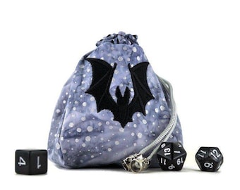 May include: A grey and silver dice bag with a black bat design. The bag has a drawstring closure and a silver metal toggle. The bag is filled with black dice with white numbers.