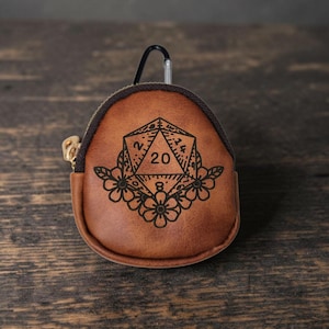 May include: A small, rounded brown leather pouch featuring a black laser-engraved design of a twenty-sided die surrounded by floral patterns. The pouch includes a dark brown zipper closure and a black metal carabiner clip attached to the top.