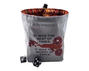 Puede incluir: Bolsa de dados de tela gris con un diseño de dragón y el texto "IT WAS THE BEST OF TIMES, IT WAS THE WORST OF TIMES". La bolsa tiene un interior estampado en naranja y marrón y un cierre de cordón. Dos dados están junto a la bolsa.
