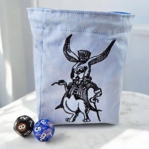 May include: A light blue fabric dice bag with a black embroidered image of a rabbit wearing a top hat and holding a cane. The bag is sitting on a white surface with two dice beside it.