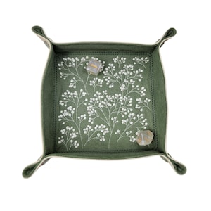 May include: A square, olive green felt tray with a white floral embroidery design. The tray has a stitched border and rolled corners. Two dice are inside the tray.