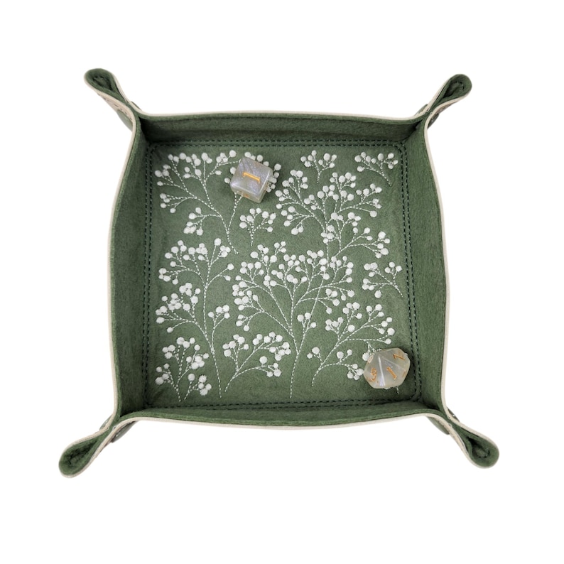 May include: A square, olive green felt tray with a white floral embroidery design. The tray has a stitched border and rolled corners. Two dice are inside the tray.