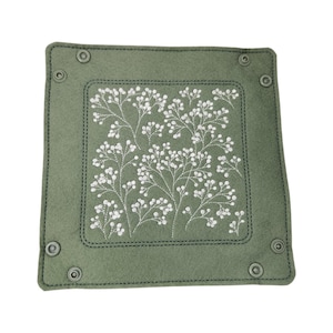 May include: A square, sage green felt tray with a white floral embroidery design. The tray has a stitched border and metal snap closures at each corner. The embroidered design features delicate white flowers and stems.