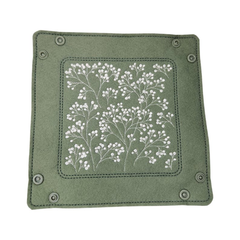 May include: A square, sage green felt tray with a white floral embroidery design. The tray has a stitched border and metal snap closures at each corner. The embroidered design features delicate white flowers and stems.