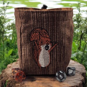 Peut inclure: Un sac à dés marron avec un écusson d'écureuil. Le sac a un aspect texturé semblable à du bois. Plusieurs dés sont éparpillés autour de la base du sac. L'arrière-plan est un cadre naturel en plein air avec de la verdure et de l'eau.