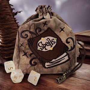 Puede incluir: Una bolsa de dados beige con un diseño de libro bordado en marrón que dice "Spells". La bolsa tiene un patrón veteado y un cordón marrón. Tres dados están al lado de la bolsa.