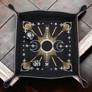 May include: Black felt dice tray with a gold and silver embroidered design of moons, stars, and sun rays. The tray has four corners with flaps that fold up to create a box.