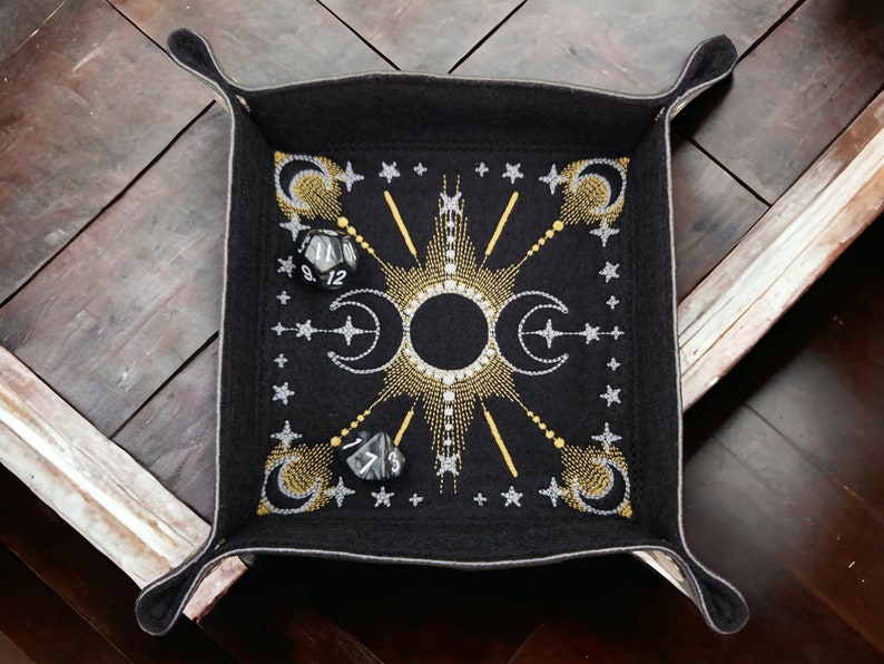 May include: Black felt dice tray with a gold and silver embroidered design of moons, stars, and sun rays. The tray has four corners with flaps that fold up to create a box.